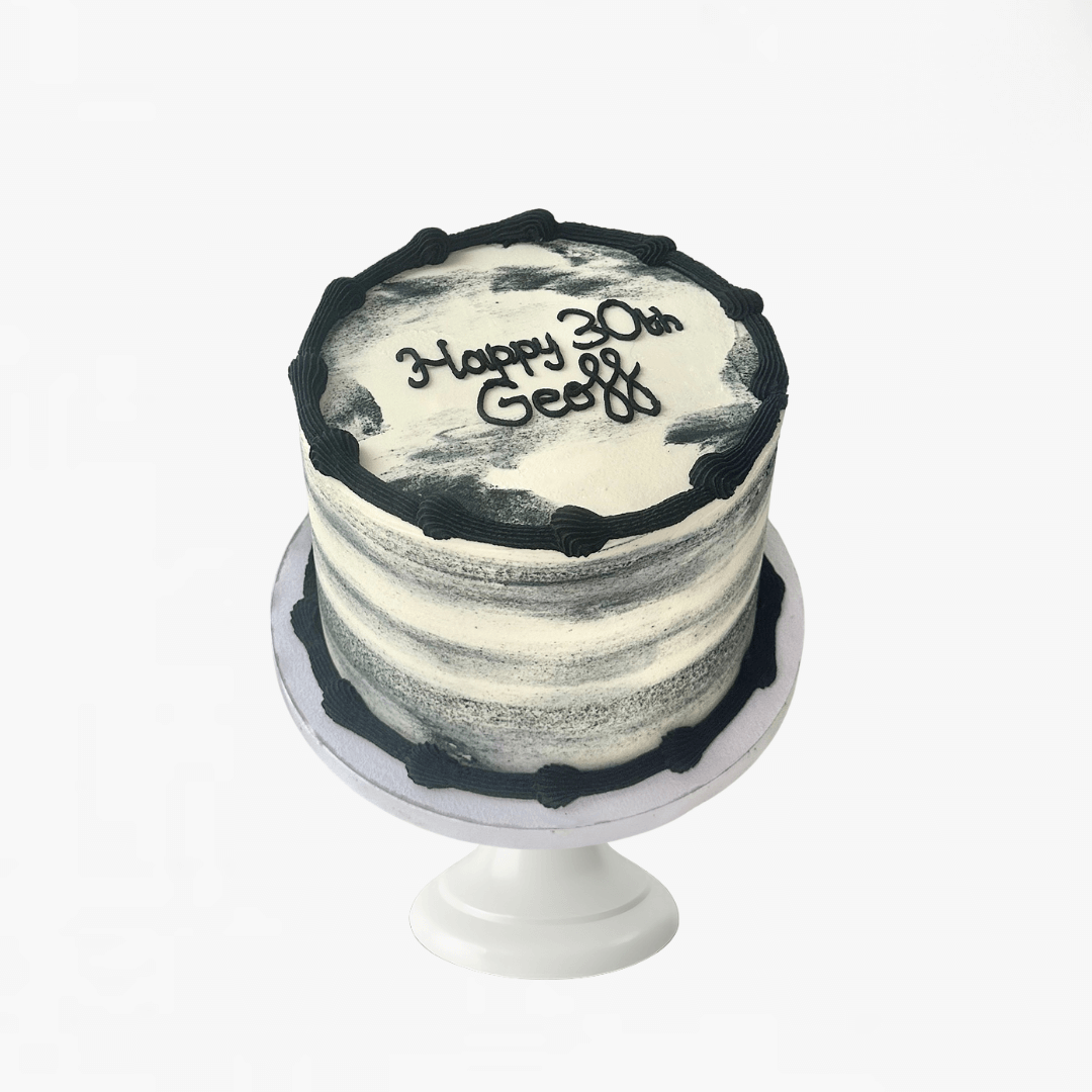 Small round cake with black and white frosting on a white stand against a white background - men's birthday cake Sydney