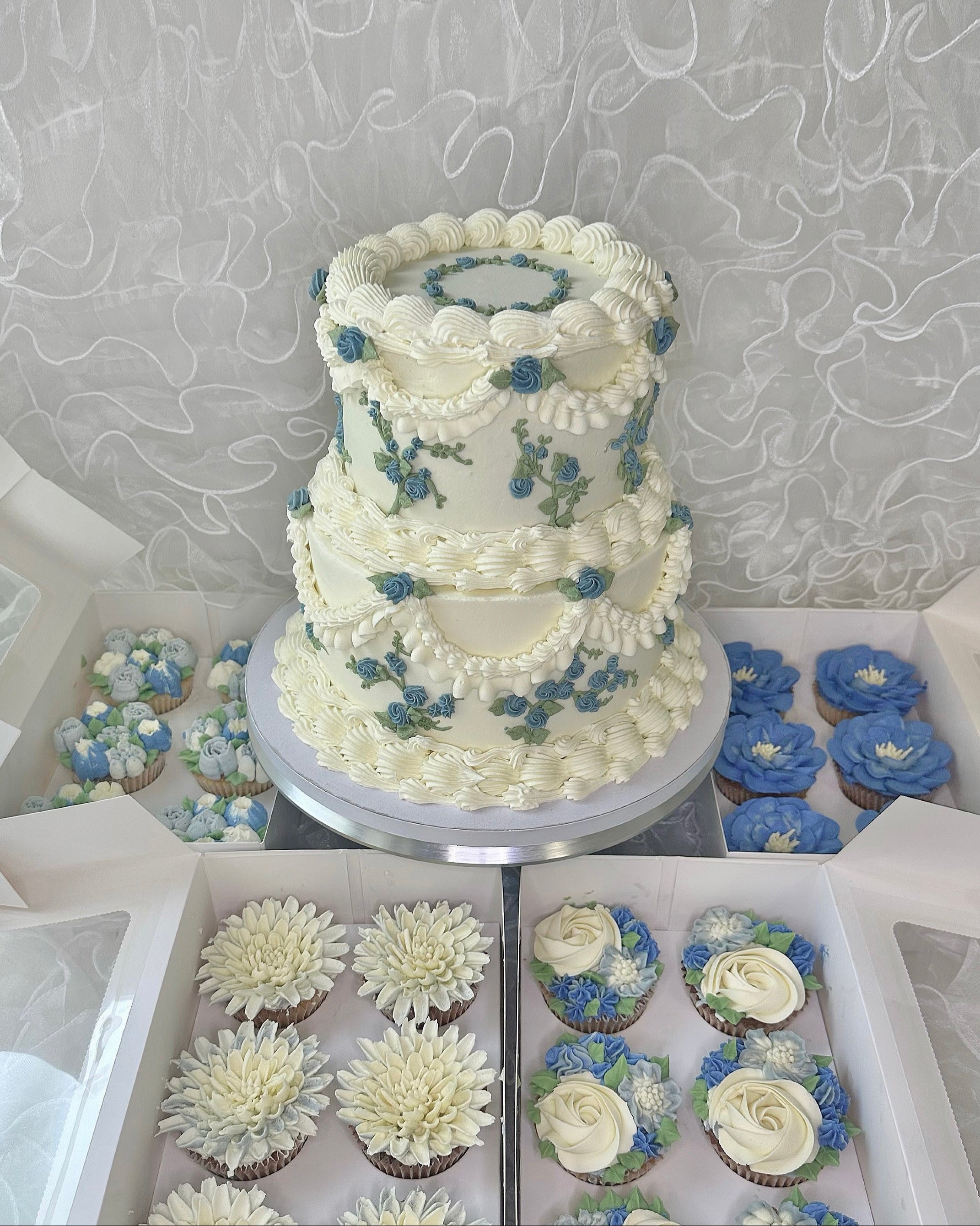 Two-tiered  cake with floral decorations surrounded by cupcakes.