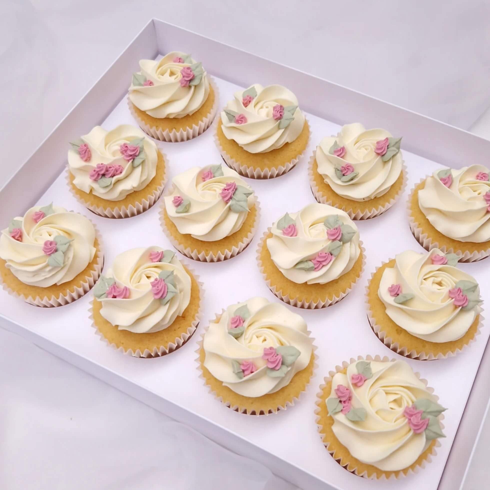 Dozen cupcakes with white frosting and decorative flowers in a box on a light background