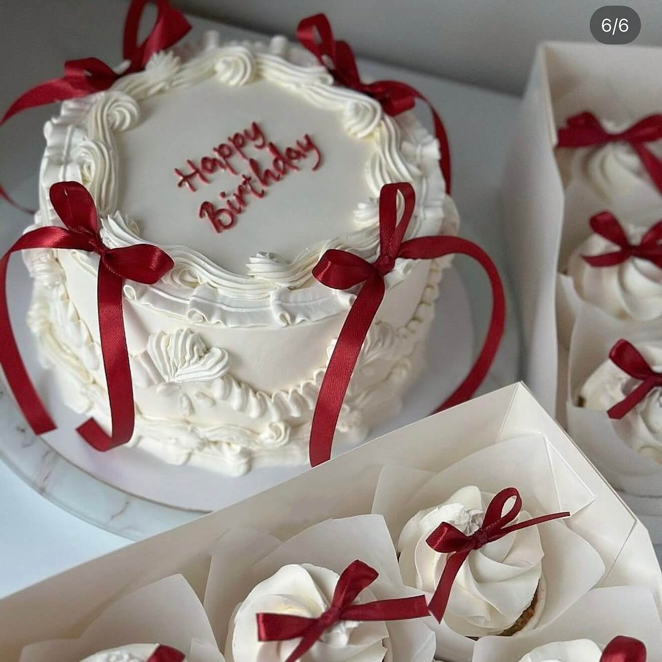 White birthday cake with red ribbons and 'Happy Birthday' text, surrounded by cupcakes.