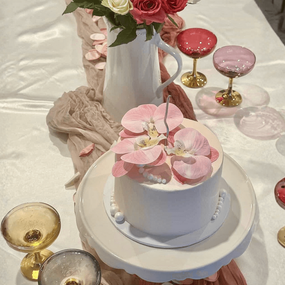 Small white cake with pink flowers on a white surface with glasses and a vase of flowers - custom cakes sydney